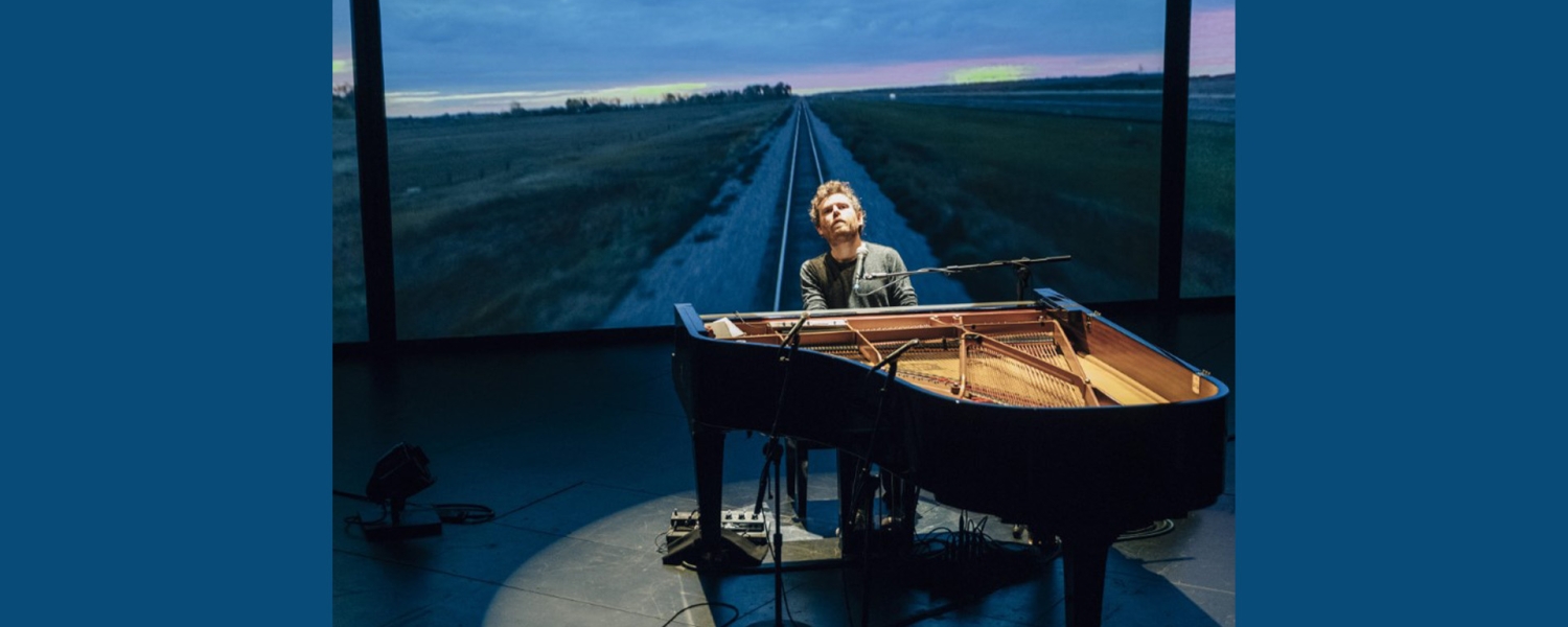 A man sits at the piano, his head tilted back, a spotlight illuminating him in a circle of light. Behind him is an image of an empty road stretching off to a sunset horizon.