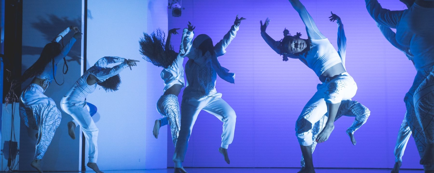 Several dancers on stage wearing white with blue and purple lighting