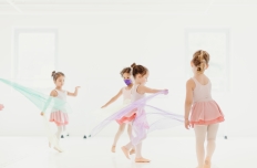four little girls in dance skirts and leotards and tights are jumping and running in a dance studio