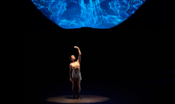 A female dancer wearing a beige tank top and shorts stands on a dark stage illuminated by the blue amorphous shape that hangs above her.
