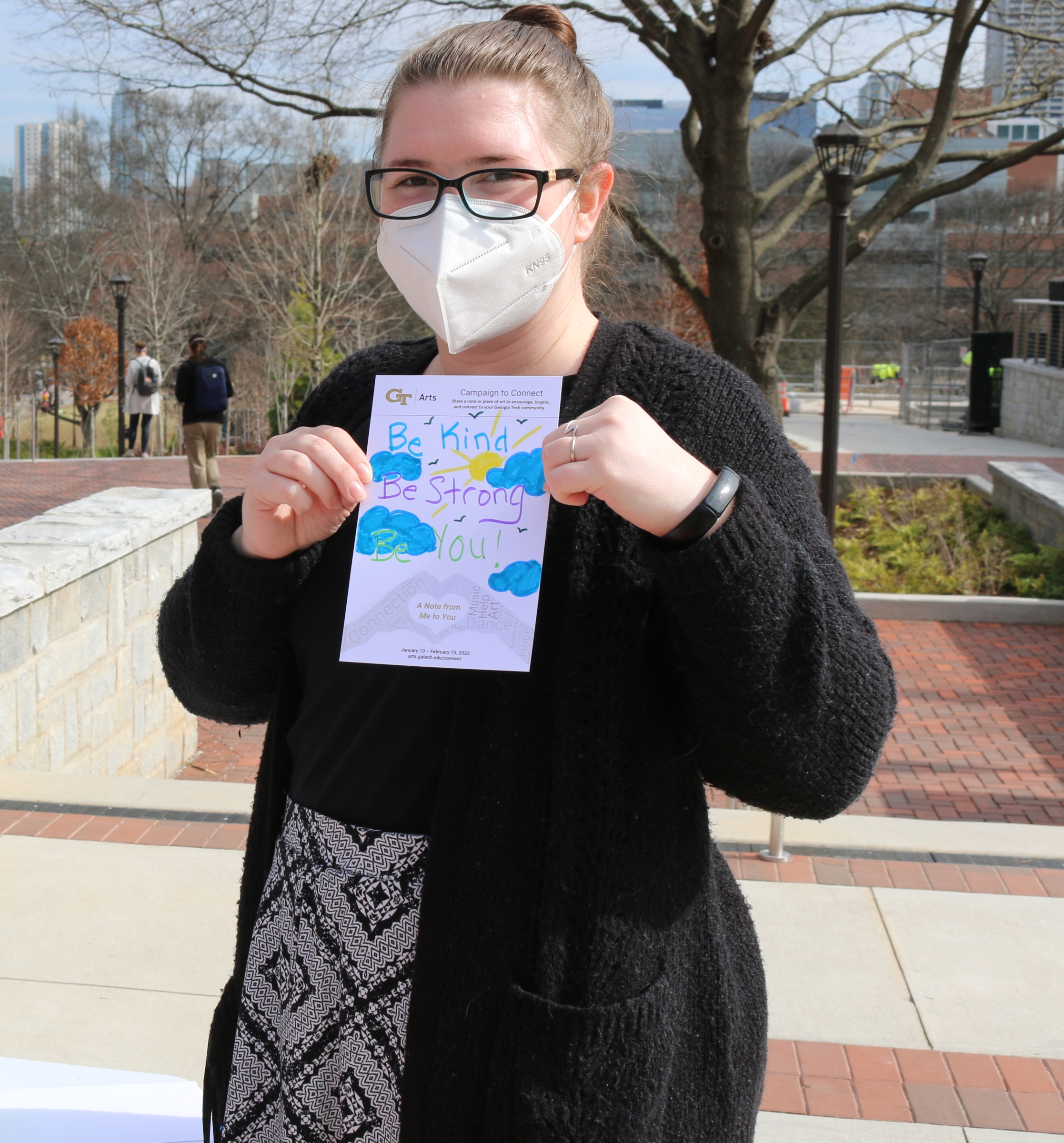 A woman stands outside holding a note card in front of her that reads Be Kind Be Strong Be You