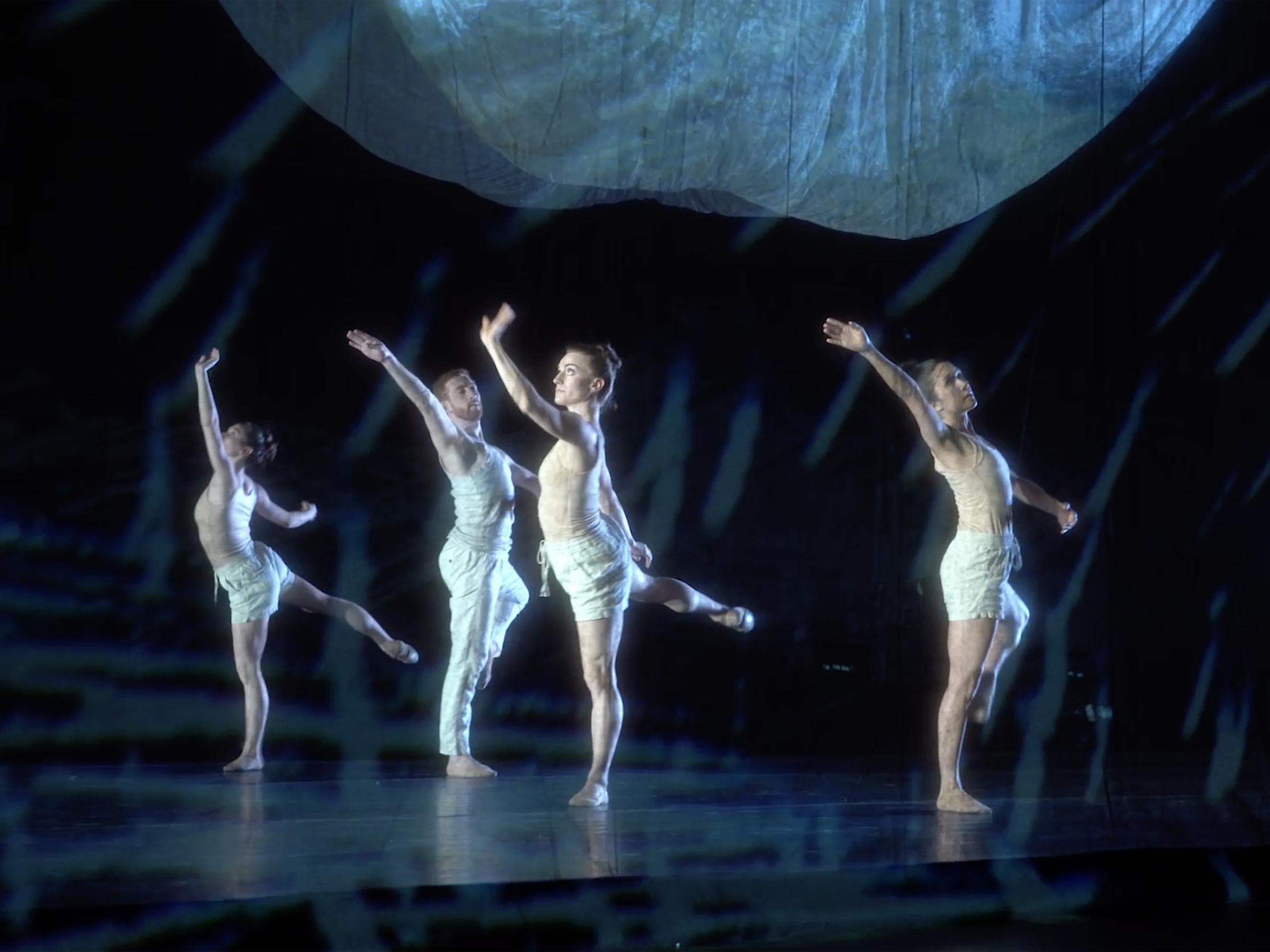 four ballet dancers on a dimly lit stage with an amorphous shape hung above them