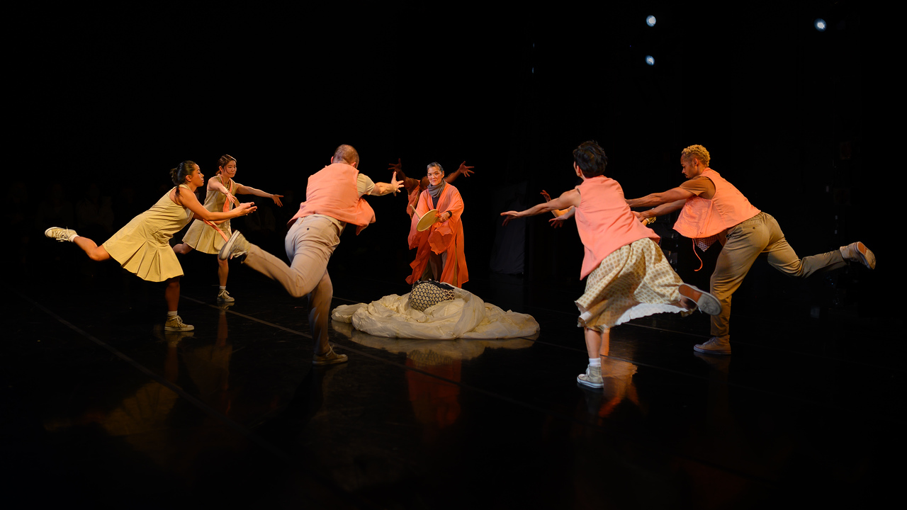 Nine dancers dressed in muted tones with bright orange touches stand on a stage in a circle surrounding a woman in orange standing in the center of a large piece of white fabric