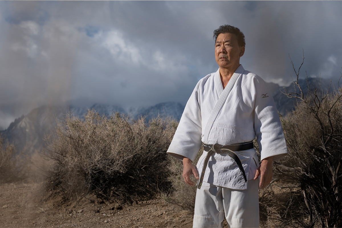 A man wearing a white kimono top and pants, standing with legs apart and arms stiffly by his side. Behind him is s rough landscape of scrub grasses