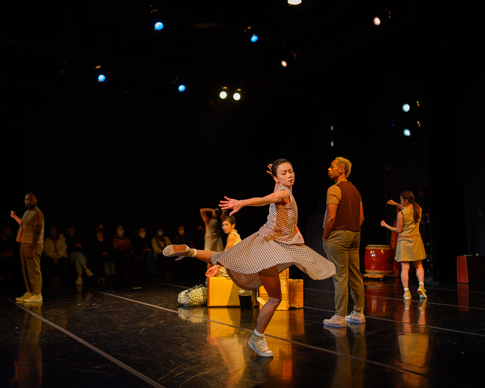 A female dancer is in mid-turn, one leg raised and bent behind her. Other dancers are standing, scattered around the stage along with old suitcases and Taiko drums.