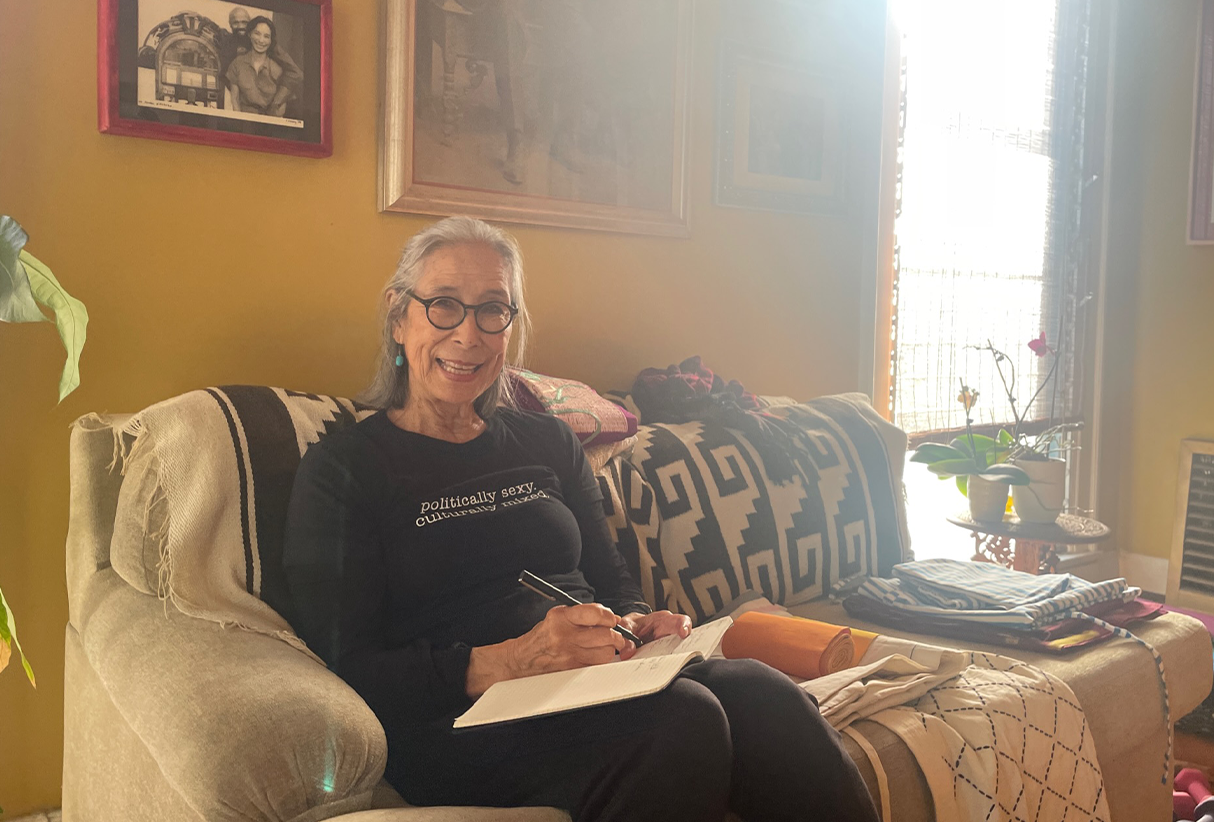 a woman with grey hair, black round eyeglasses and a big smile sits on a couch with a notebook and pen in her hand