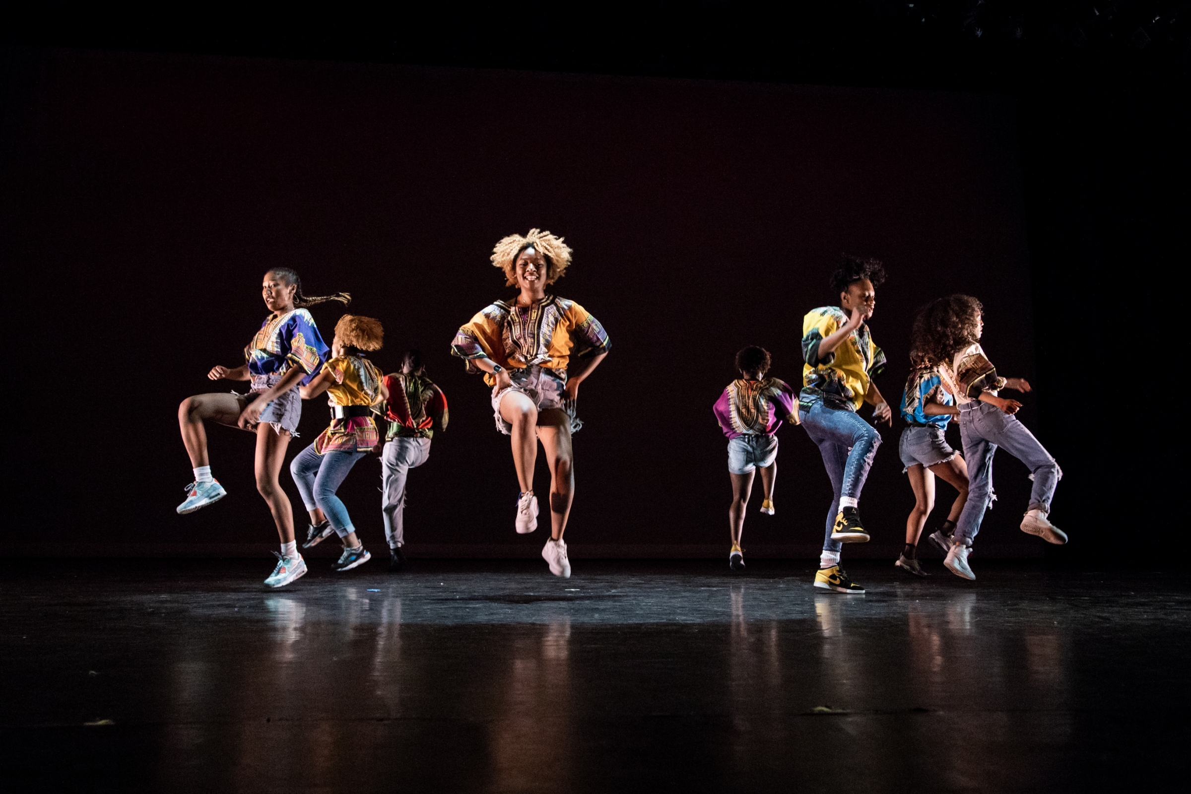 The women of LDC performing for the annual Ladies of Hip - Hop festival, wearing multi - color dashikis, faced outward, in a circle in front of a background