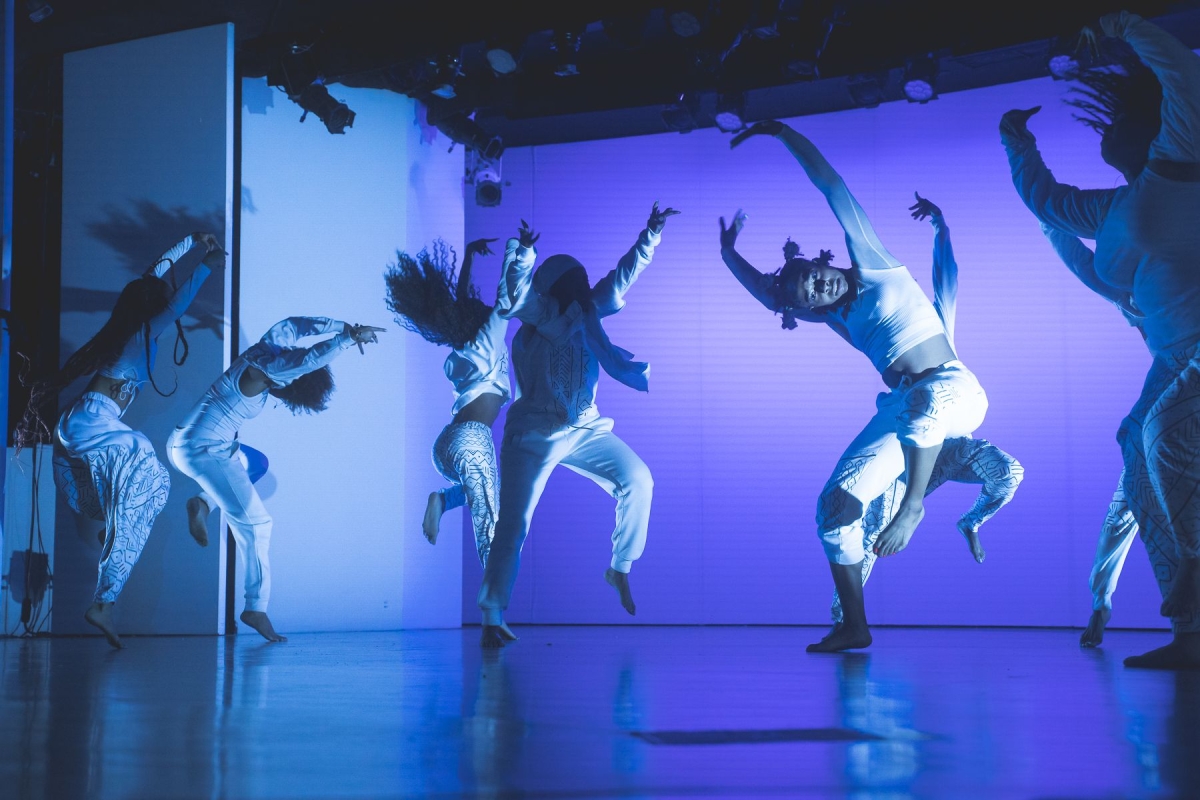 Several dancers on stage wearing white clothing with purple and blue environment lighting