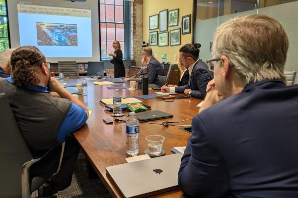 People sitting around a conference table listen to a presentation. A presentation slide reads, "Technology creates possibilities. The liberal arts turns those possibilities into impact."