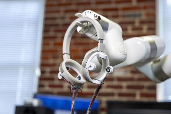 Robotic arm holding circular sensor devices with exposed wiring in a lab setting with a blurred brick wall background.