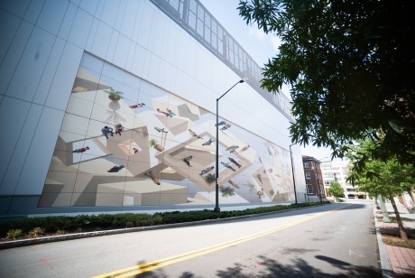 A mural is painted onto the side of a 21-story building. The mural is 150 feet wide and 80 feet high. A jumble of shapes, all with right angle, are populated with small human figures variously at rest, standing, or sitting.