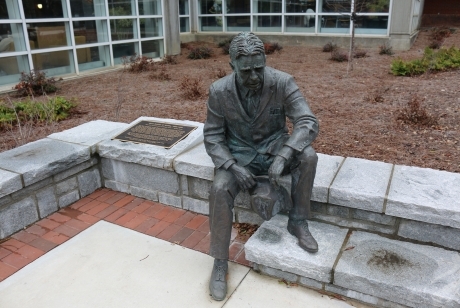 A darkened bronze statue of a seated man, dressed in a business suit. His hat is in his hands. He is sitting on a low granite wall, his right foot on the ground and his left foot perched higher up on a low step.