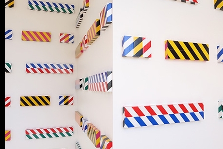 Three white walls, at 90 degree angles to each other, are hung with more than 30 different flat, rectangular canvas each covered in different combinations of industrial safety tape.