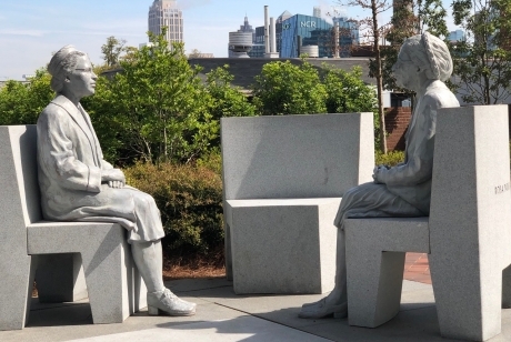 Figures of Rosa Parks at age 42 and age 92, seated on separate chairs facing eachother with an empty chair between them.
