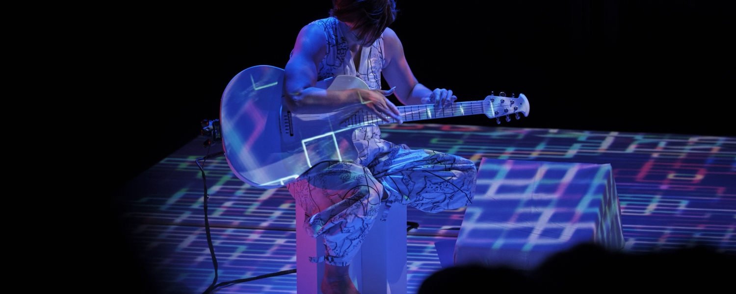 Against a black background, a woman in a silver jumpsuit sits on a chair playing a white guitar. Digital patterns in blue, purple and white are projected on her, the guitar, and the floor around her.