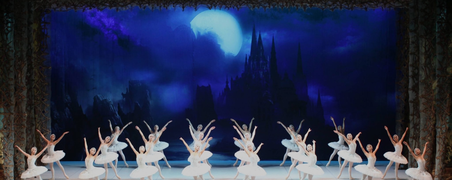 Ballerinas in white tutus are en pointe, their arms above their heads with hands crossed. The prima ballerina is in front with two perfectly straight lines of ballerinas behind her. The scene is blue, as a lake.