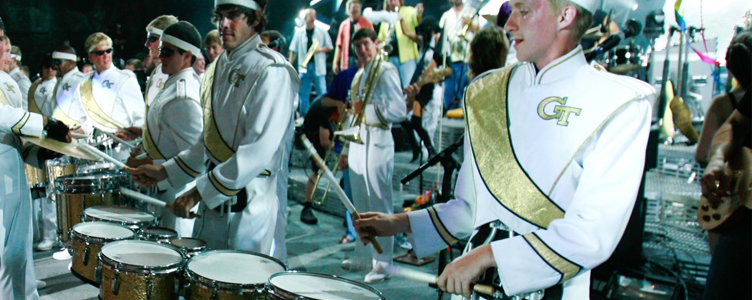 the drum section of a marching band. College students dressed in white uniforms with gold sashes and the GT logo, and a white headband.