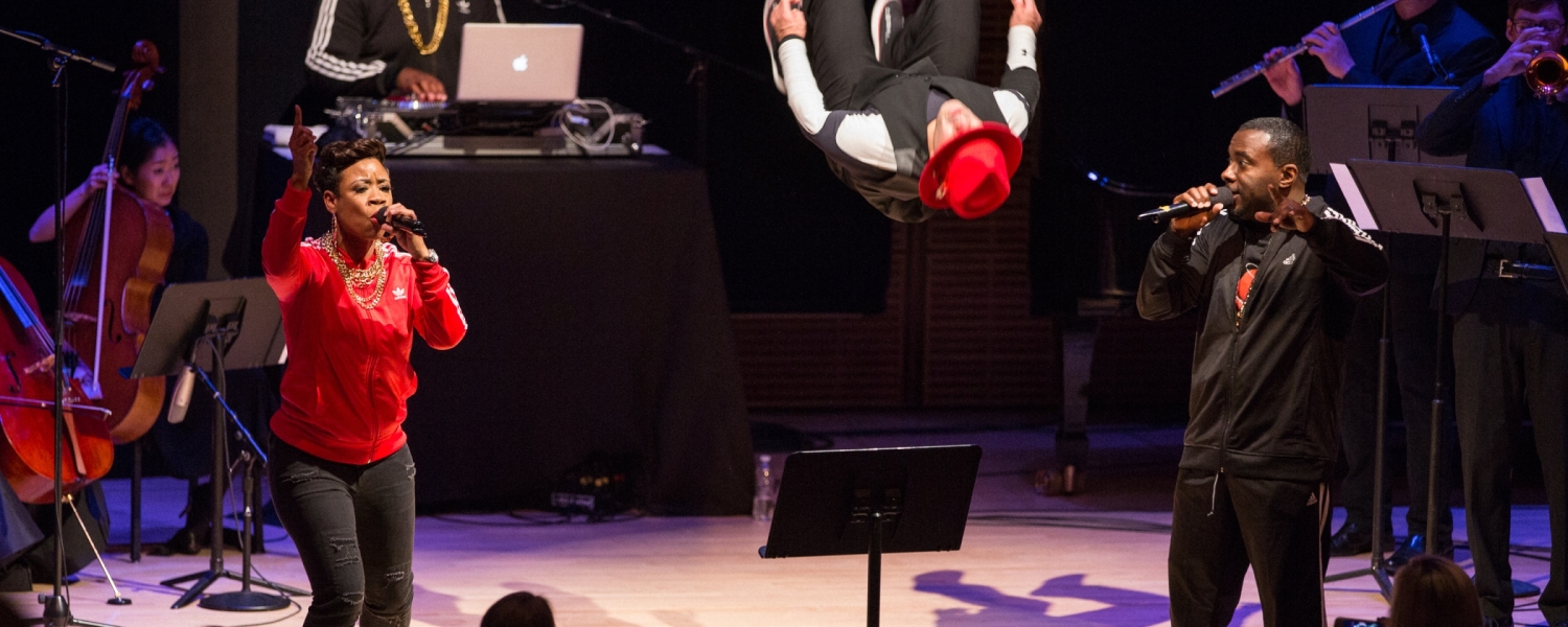 Singers, musicians, and dancers are on stage. The dancer is mid-flip, his red-capped head seeming to float four feet off the ground.
