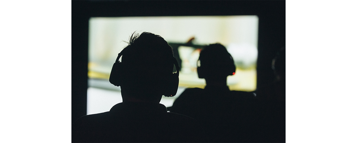 The silhouette of two people seen from the back, each wearing headphones and looking at a projected image on a movie screen. 