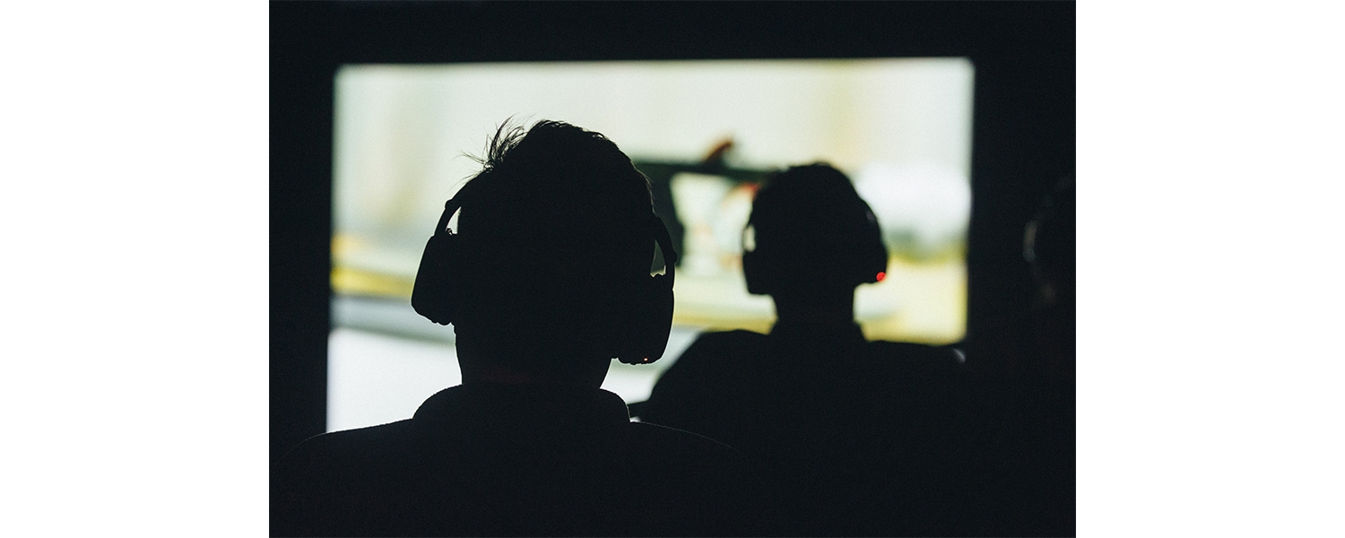 The silhouette of two people seen from the back, each wearing headphones and looking at a projected image on a movie screen.