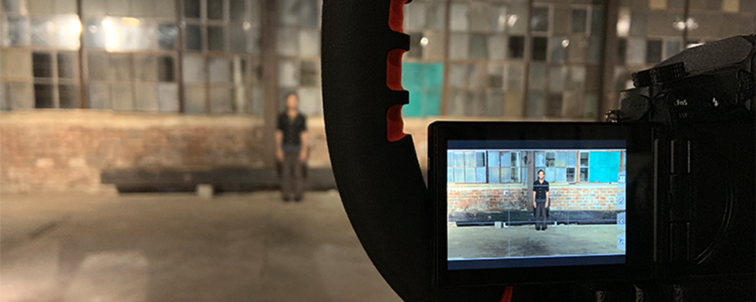 In the foreground we are looking at a small monitor of a video camera, capturing the image of a person standing 30 feet away. Blurry in the far distance of the photo itself is a person standing on a cement floor in front of a high wall of small, square windows.