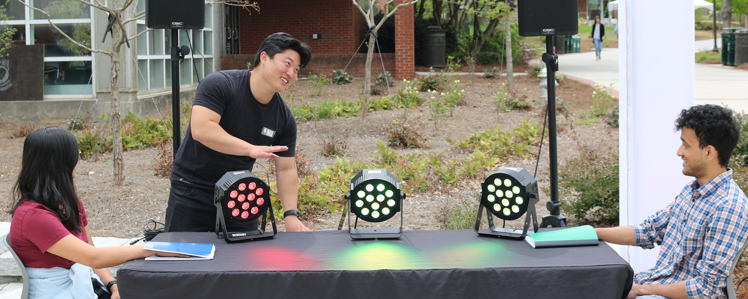 A young man stands in front of a table with sensors and colorful lights on top, while a young woman and man sit at opposite ends smiling at him. 