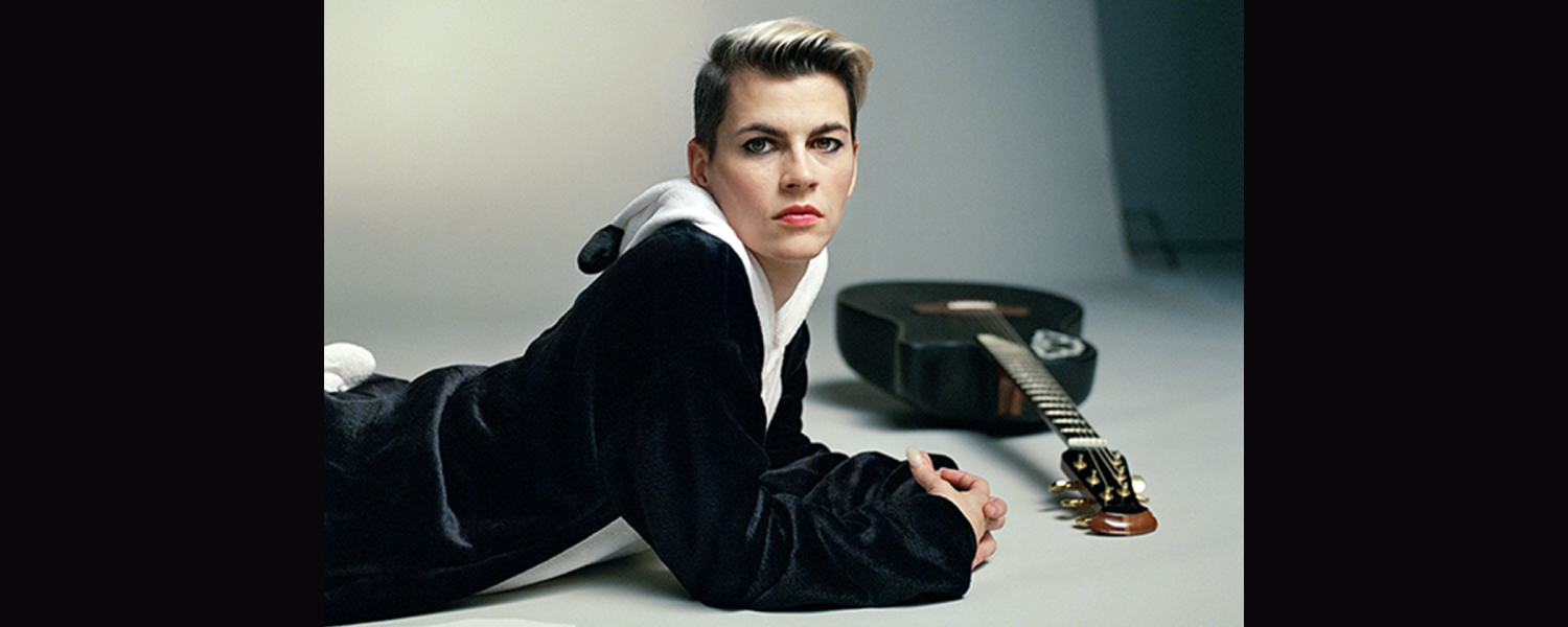 Kaki King wearing a black and white panda jumpsuit lies on the ground, her black guitar on the floor beside her.