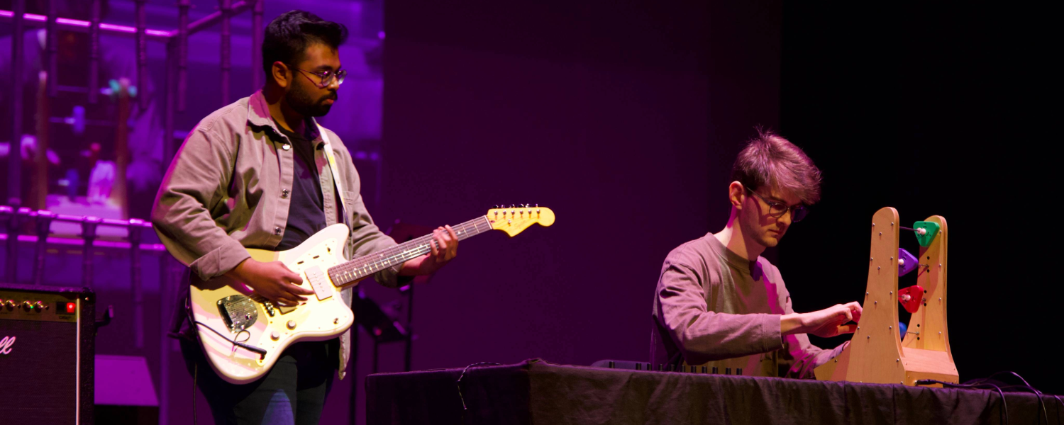 One person standing holding guitar, second person sitting using instrument