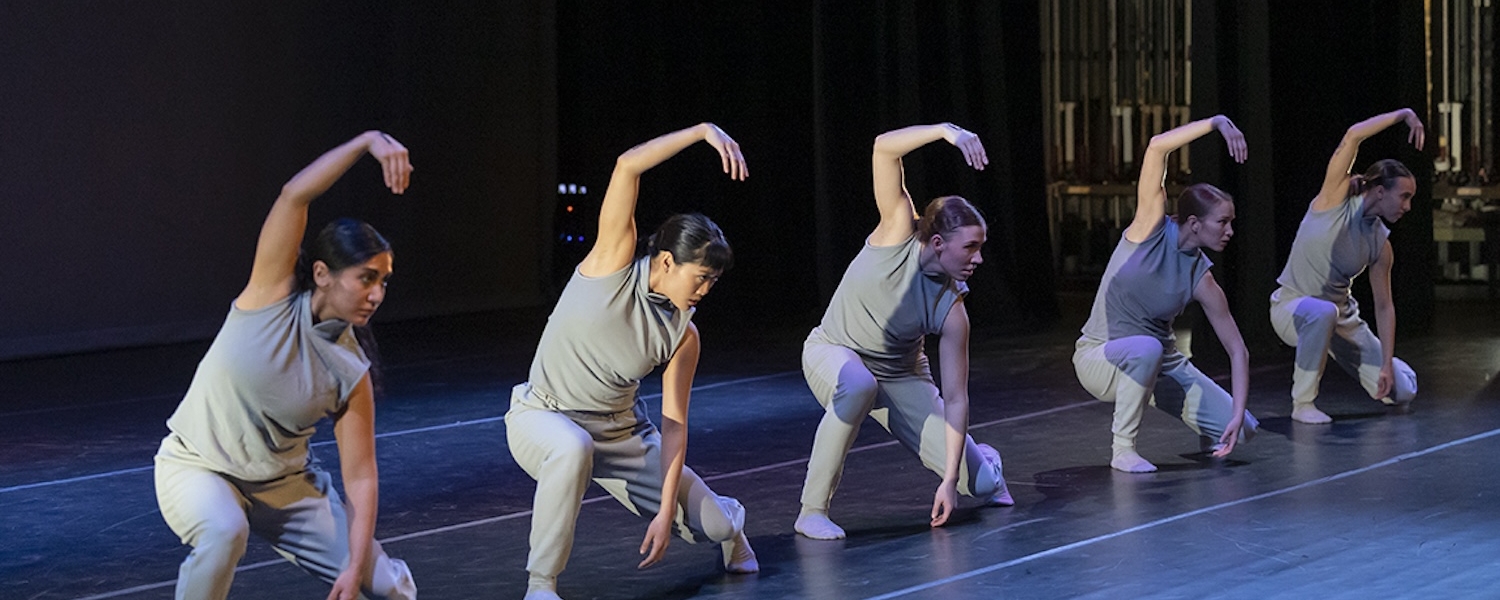 five dancers on stage wearing grey shirt and pants with one arm above head and other arm towards ground