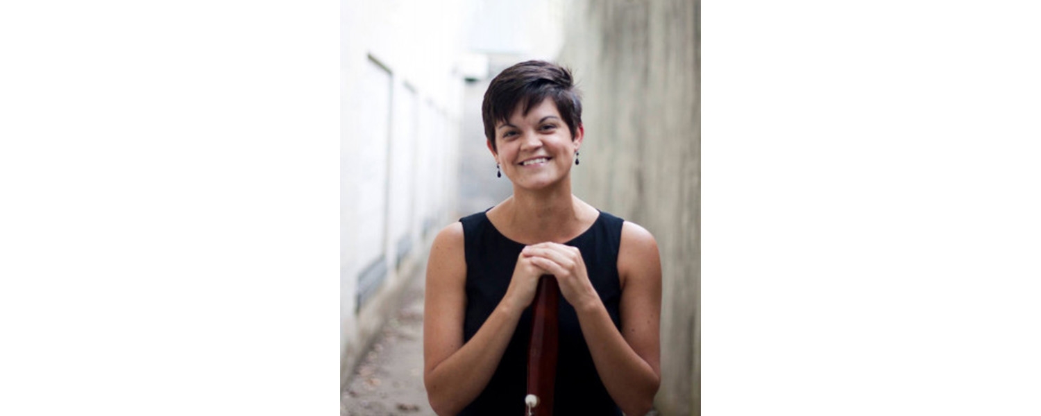 person with short brown hair wearing black sleeveless top standing with hands propped on instrument and smiling at camera