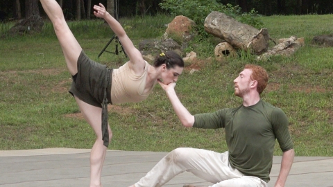 The setting is an outdoors, the woods and grass surrounding a low stage. A male dancer sits, one hand touching the ground, the other gently cupping the female dancer's face. She is posed in a deep arabesque towards the man, her face in his hand and her upper arm and leg extended skyward.