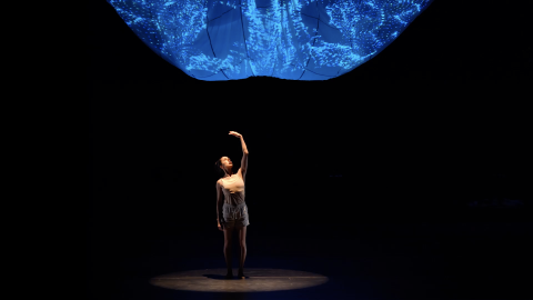 A woman wearing beige shorts and tank top stands alone in the spotlight, her left arm raised above her head as she looks towards her palm. Above her looms an amorphous blue image.