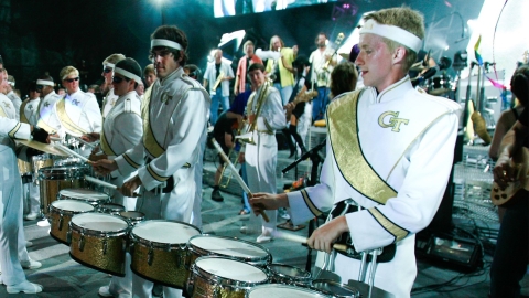 the drum section of a marching band. College students dressed in white uniforms with gold sashes and the GT logo, and a white headband.