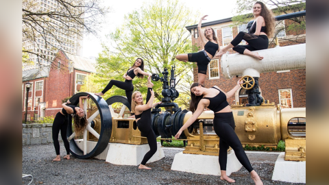 Six white women in black crop tops and leggings are in various dance poses on top of and besides the gold and white Georgia Tech steam engine.