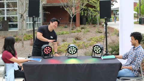 A young man stands in front of a table with sensors and colorful lights on top, while a young woman and man sit at opposite ends smiling at him. 