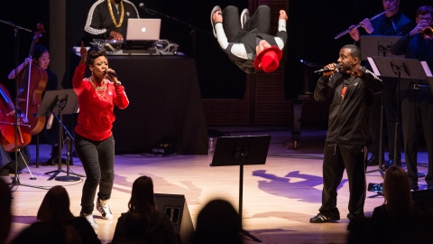 Multiple people on stage wearing black, red or white, with microphones, instruments, and laptop
