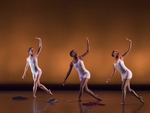 three ballerinas each wearing close fitting pale tank tops and shorts, are standing with their front leg bent, other leg slightly raised to the back, one arm raised to turn their torsos towards the camera