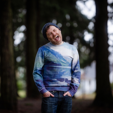 A man wearing blue jeans and a long sleeve Tshirt printed in abstract blues stands with his hands in his front pockets. He is fully in focus, his eyes closed and his mouth open in song; trees are blurry behind him.
