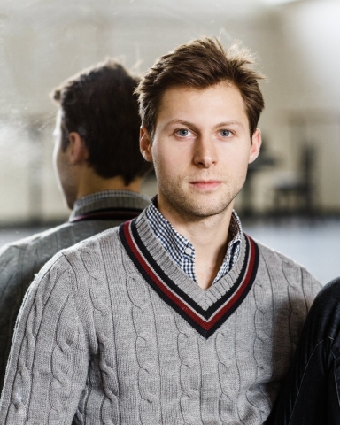 A man wearing a grey sweater over a blue checked shirt. He is facing the camera but his back is to a dance studio mirror so we see both sides of him.