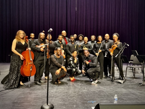 A group of approximately 20 musicians all dressed in black holding classical music instruments