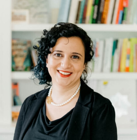 A woman with short curly black hair smiles at the camera. She is wearing a pearl necklace, a black suit jacket and black shirt, and red lipstick. The background behind her are of white bookshelves with different books of varying bright colors.