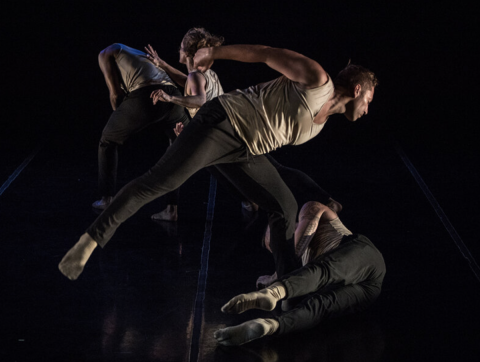 Four dancers wearing loose tan tank tops and black pants perform on a dark stage. We cannot clearly see their faces. From left to right: A male dancer with his back hunched away from a female dancer reaching towards him. In front of both are another pair of dancers, the male dancer leaning on his left foot and looking down at the female dancer, who is on laying next to his left leg, on her right side on the floor, her legs extended forward.