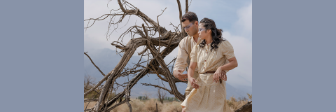 Two figures facing the camera but looking downward to the side, the man behind the woman with his arms around her waist. Twisted, gnarled, bare branches are next to them. Behind them is a range of mountains and smoky blue sky and clouds.