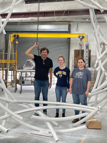 A man and two women are standing in a large, bright studio surrounded by heavy equipment and a circular sculpture