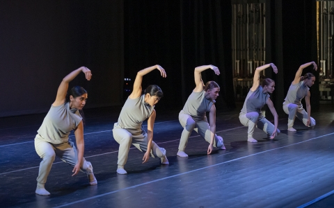five dancers on stage wearing light grey shirt and pants with one arm curved above their heads and and body lowered toward ground