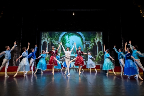 group of ballet dancers on stage wearing blue and red attire with garden scene displayed in background