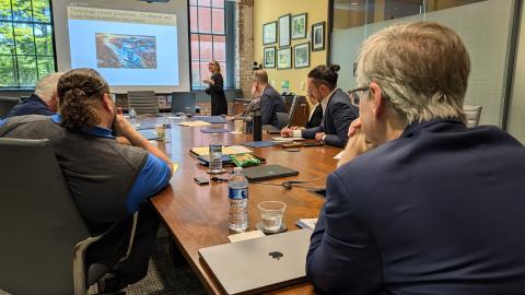 People sitting around a conference table listen to a presentation. A presentation slide reads, "Technology creates possibilities. The liberal arts turns those possibilities into impact."