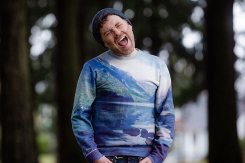 A man wearing blue jeans and a long sleeve Tshirt printed in abstract blues stands with his hands in his front pockets. He is fully in focus, his eyes closed and his mouth open in song; trees are blurry behind him.