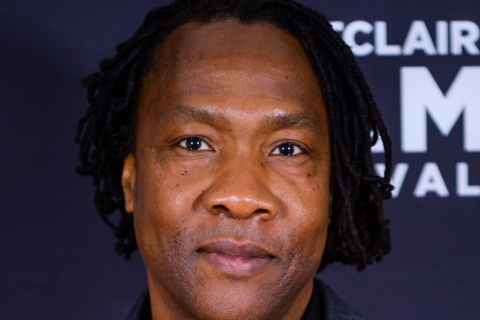 A Black man, seen from the shoulders up, hs hair is in ear-length dreadlocks, parted to the side, and he is looking directly at the camera