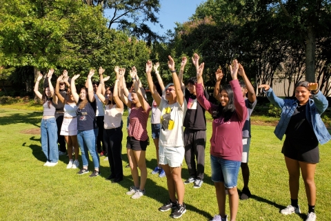 Outside on a green lawn on a sunny day, a group of approximately 20 young people stand in two lines, waving their hands in the air.